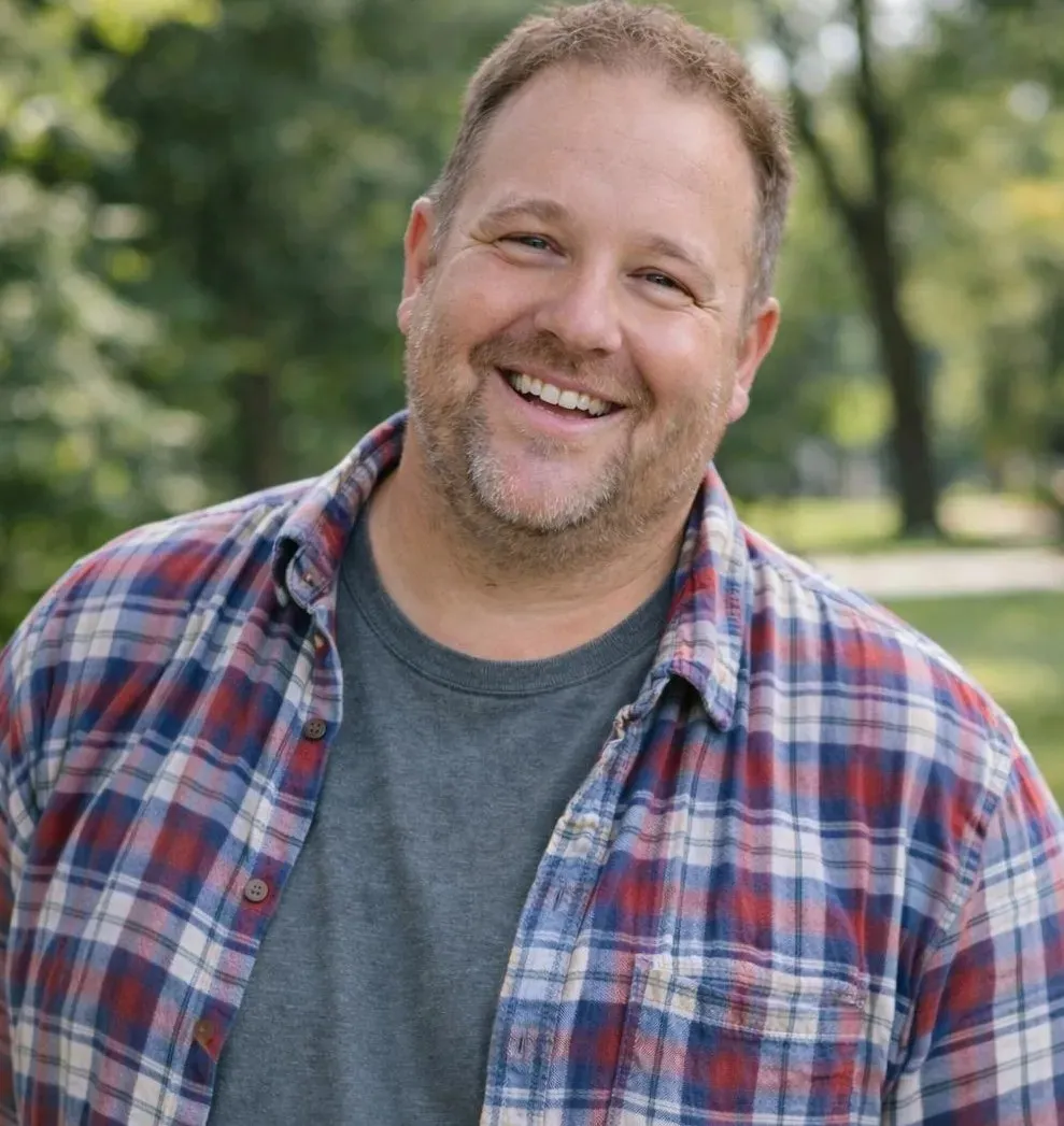 'An outdoor portrait of a smiling, middle-aged man with short brown hair and light facial stubble, wearing a red and blue plaid flannel shirt over a grey t-shirt.'