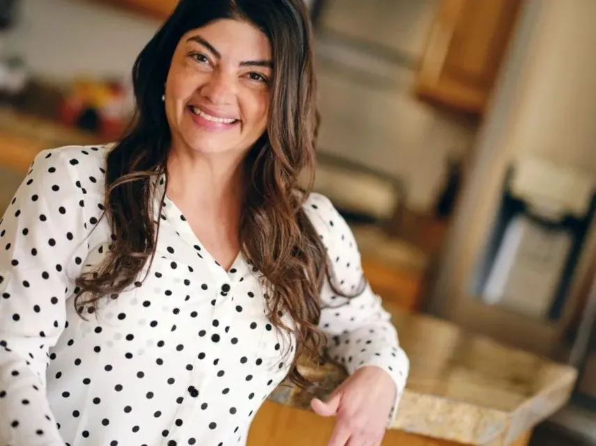 'A smiling woman with long brown hair wearing a white blouse with black polka dots, leaning against a kitchen counter.'